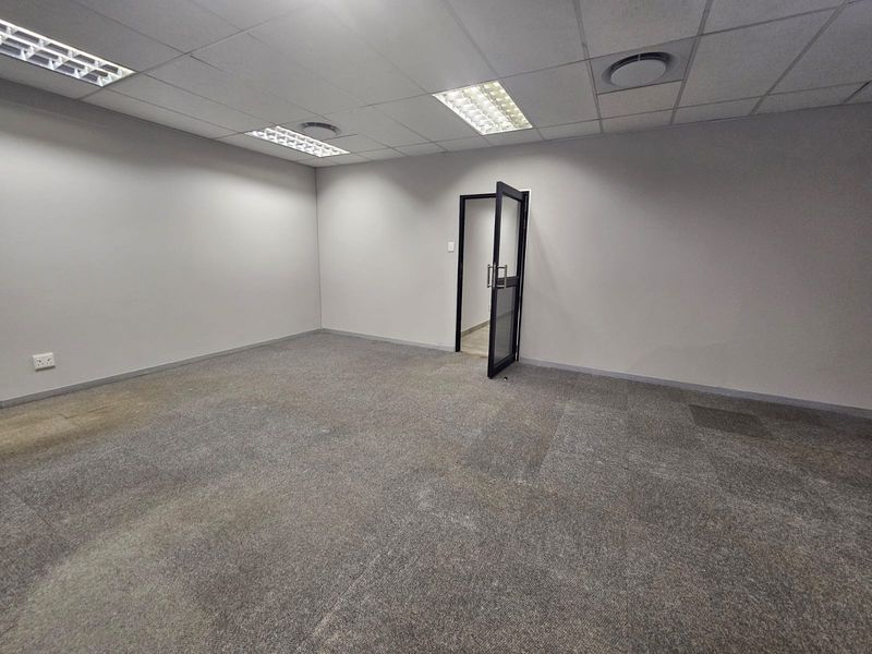 A vacant, open-plan office space featuring neutral walls, carpeted flooring, suspended ceiling with recessed lighting, and a modern glass-panelled door.