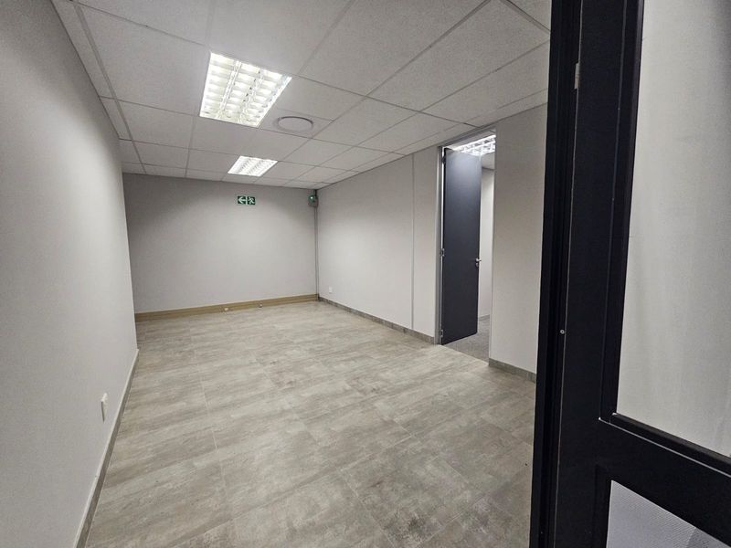 A clean, empty office space featuring neutral tiled flooring, a suspended ceiling with recessed lighting, and a clear internal doorway.