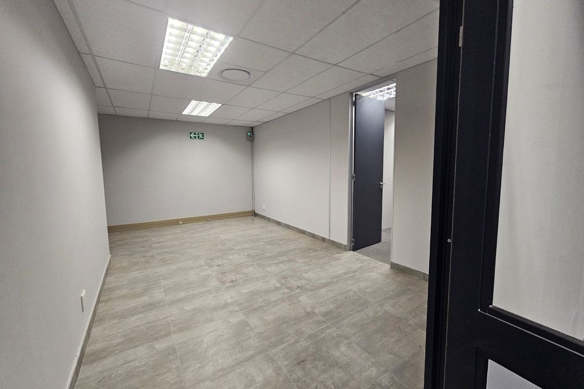 A clean, empty office space featuring neutral tiled flooring, a suspended ceiling with recessed lighting, and a clear internal doorway.