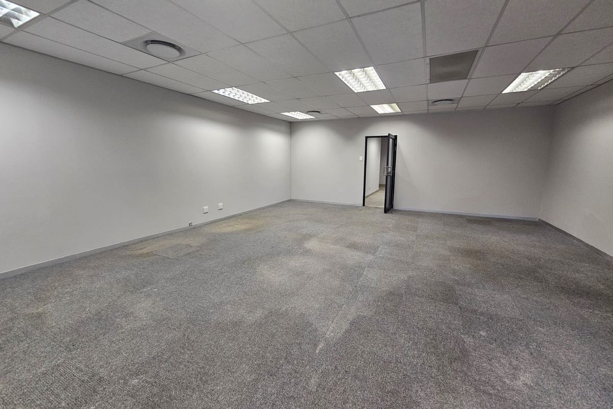 An empty, open-plan office space featuring a suspended ceiling with recessed lighting and grey carpet tiles.
