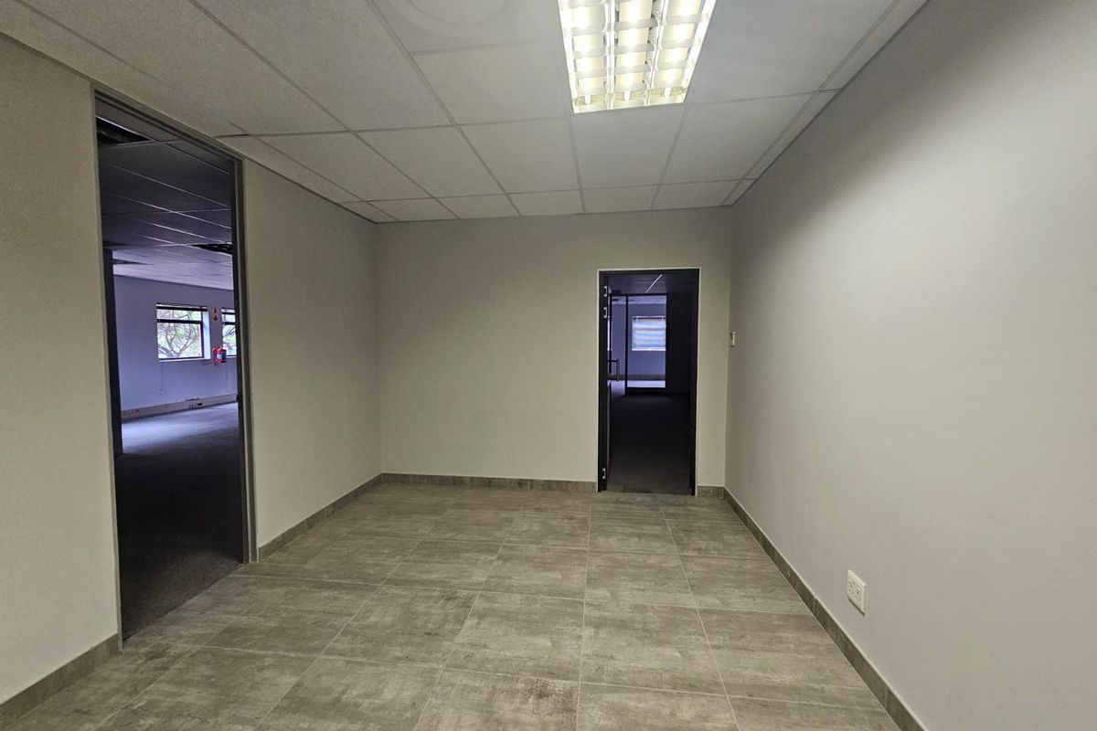 A clean, empty office space featuring neutral tiled flooring, a suspended ceiling with integrated lighting, and access to adjacent rooms.