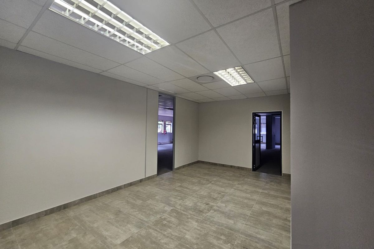 A clean, empty office space featuring neutral grey walls, modern tiled flooring, and a suspended ceiling with integrated fluorescent lighting.