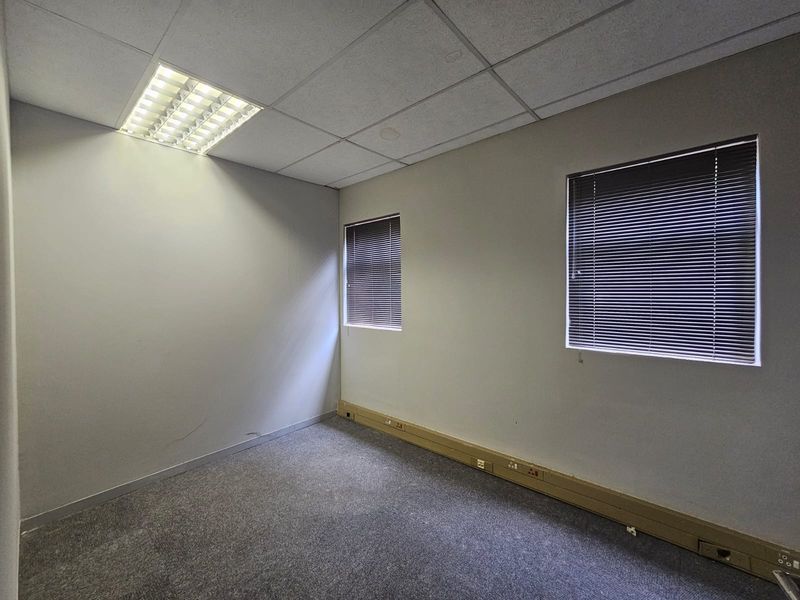 An empty office space featuring carpeted flooring, suspended ceiling with recessed lighting, and windows fitted with venetian blinds.