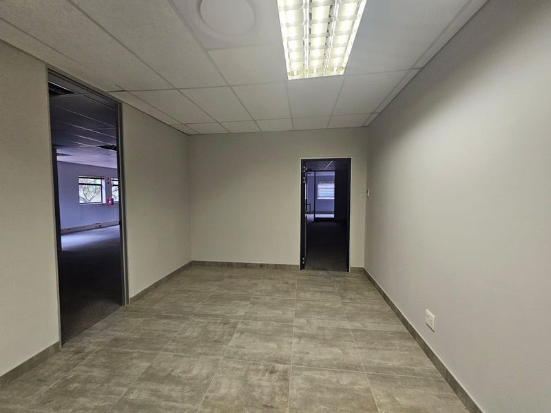 A clean, empty office space featuring neutral tiled flooring, a suspended ceiling with integrated lighting, and access to adjacent rooms.