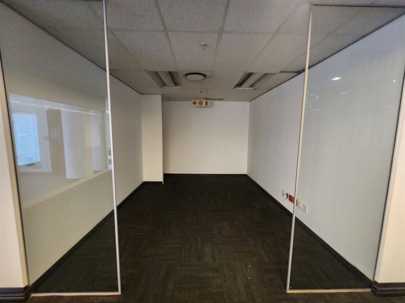 A vacant, modern office space featuring glass partition walls, carpeted flooring, and a suspended ceiling with recessed lighting.