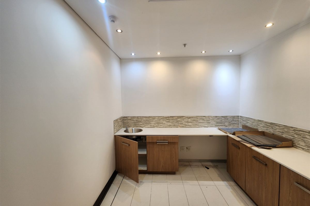 A compact office kitchenette featuring wood-finish cabinetry, a stainless steel sink, tiled backsplash, and recessed ceiling lighting.