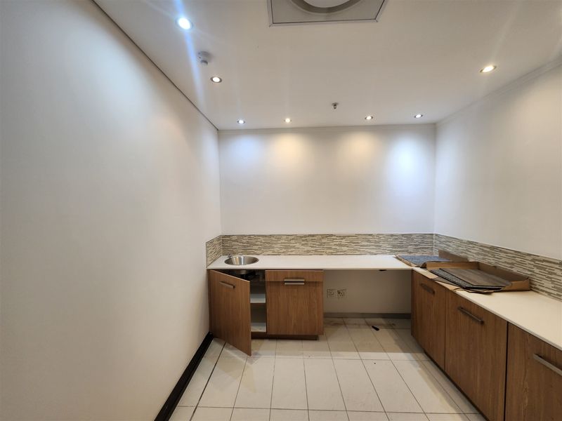 A compact office kitchenette featuring wood-finish cabinetry, a stainless steel sink, tiled backsplash, and recessed ceiling lighting.
