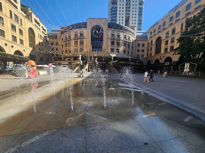 A vibrant commercial plaza featuring a central fountain, paved pedestrian area, and surrounding multi-story office and retail building architecture.
