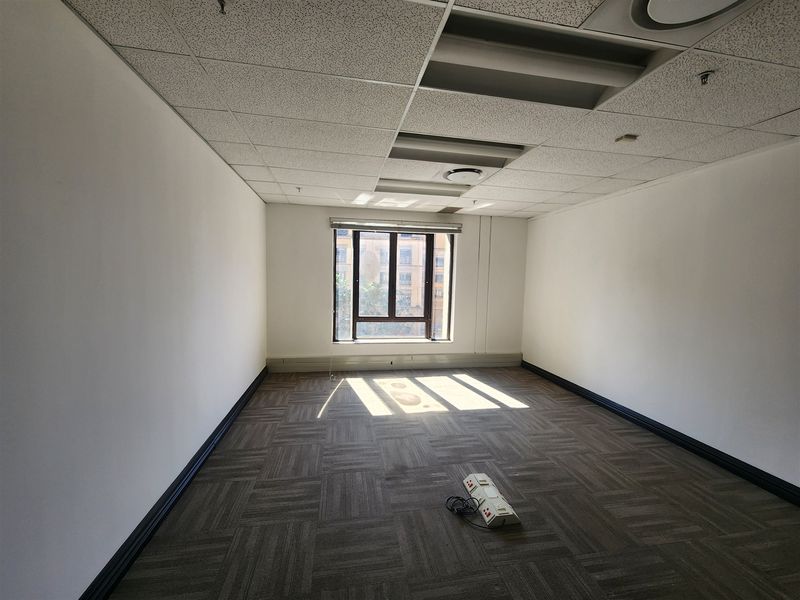 A vacant office space featuring carpeted flooring, a suspended ceiling with recessed lighting, and a window providing natural light.