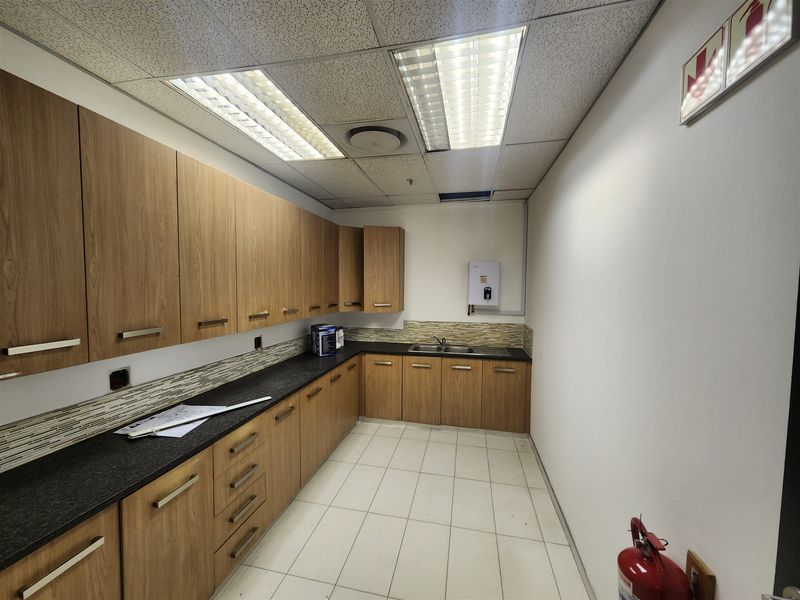 A functional office kitchenette featuring wood-look cabinetry, dark stone countertops, a double basin sink, and tiled backsplash with integrated water heating.