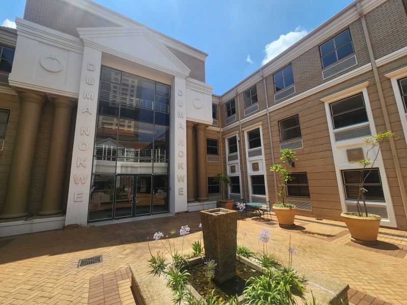 A well-maintained office building entrance featuring a brick facade, glass curtain wall, decorative columns, and a paved courtyard with a central water feature.