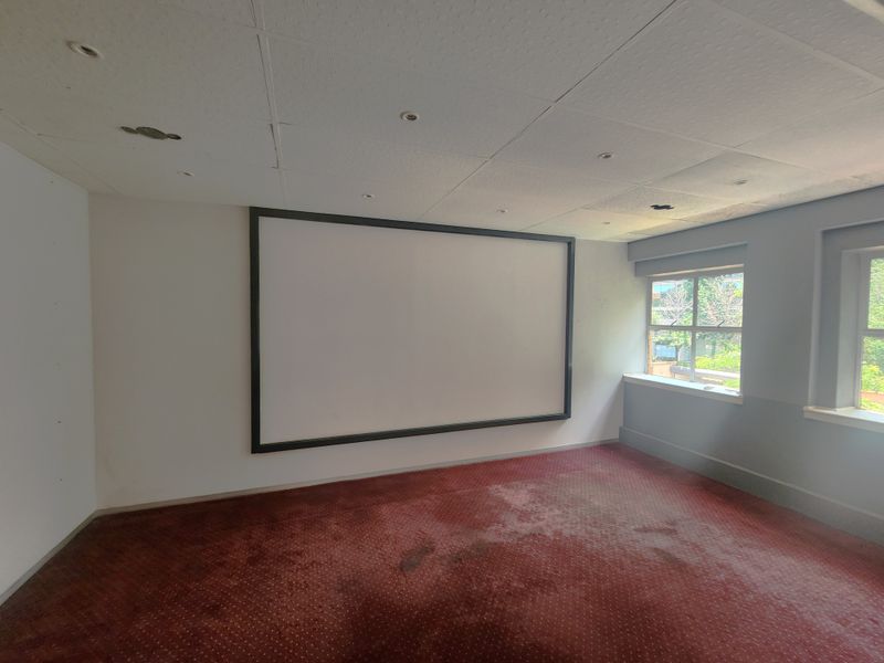 An empty office space featuring a fixed projection screen, drop ceiling with recessed lighting, and worn carpet flooring.