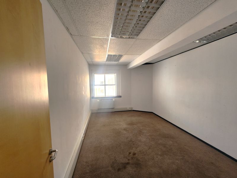 An empty office space featuring a suspended ceiling, fluorescent lighting, a window-mounted air conditioning unit, and carpeted flooring.