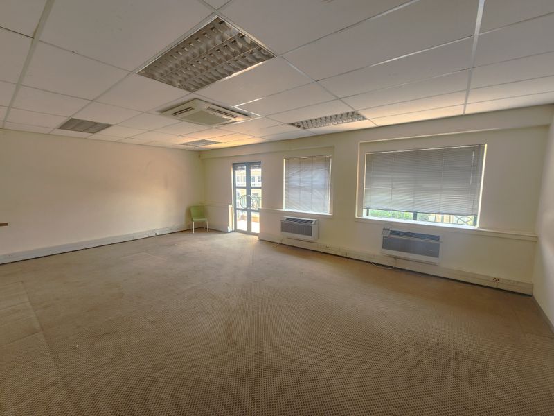 Spacious open-plan office area featuring a suspended ceiling, wall-mounted air conditioning units, and large windows with blinds.