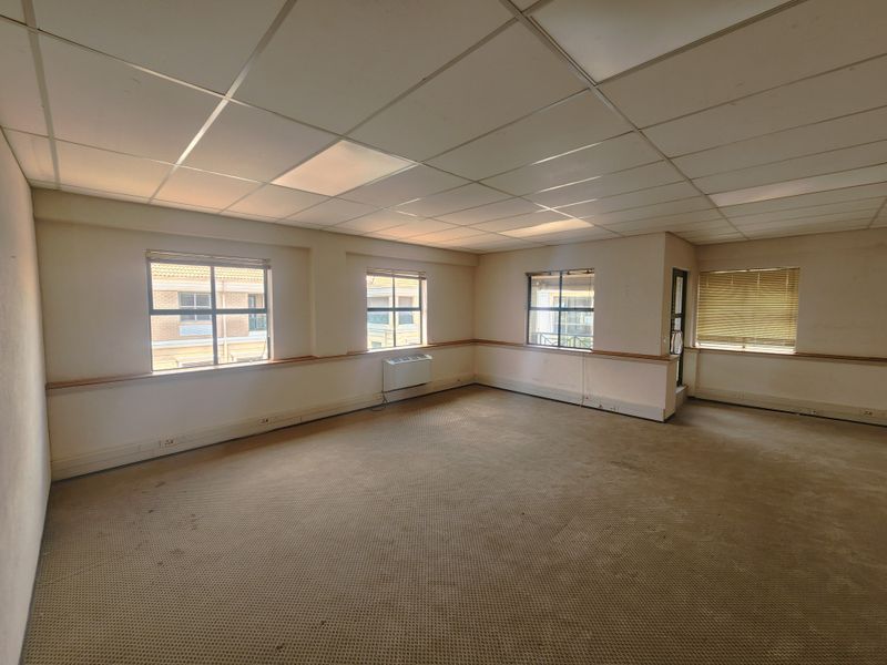 An empty, open-plan office space featuring a suspended ceiling, recessed lighting, wall-mounted air conditioning, and large windows with blinds.