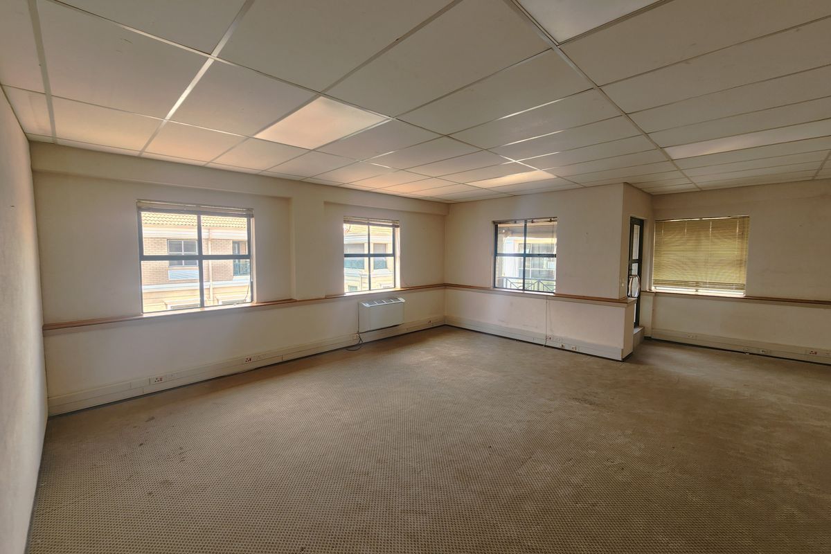 An empty, open-plan office space featuring a suspended ceiling, recessed lighting, wall-mounted air conditioning, and large windows with blinds.