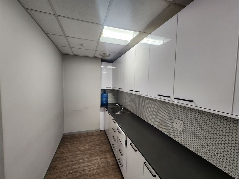 A clean, modern office kitchenette featuring white high-gloss cabinetry, a dark countertop, stainless steel sink, and a stylish hexagonal tile backsplash.