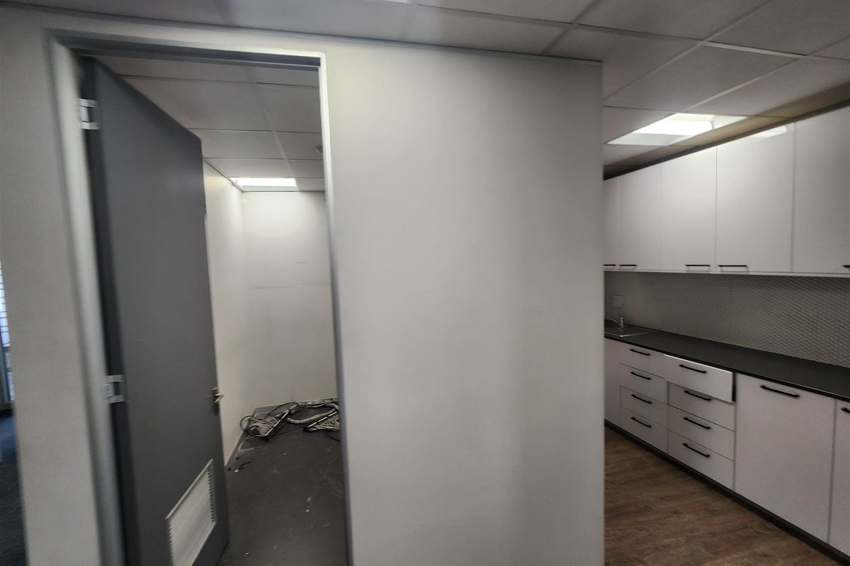 A functional office kitchenette featuring white cabinetry, dark countertops, and a drop ceiling, adjacent to an empty utility space with exposed wiring.
