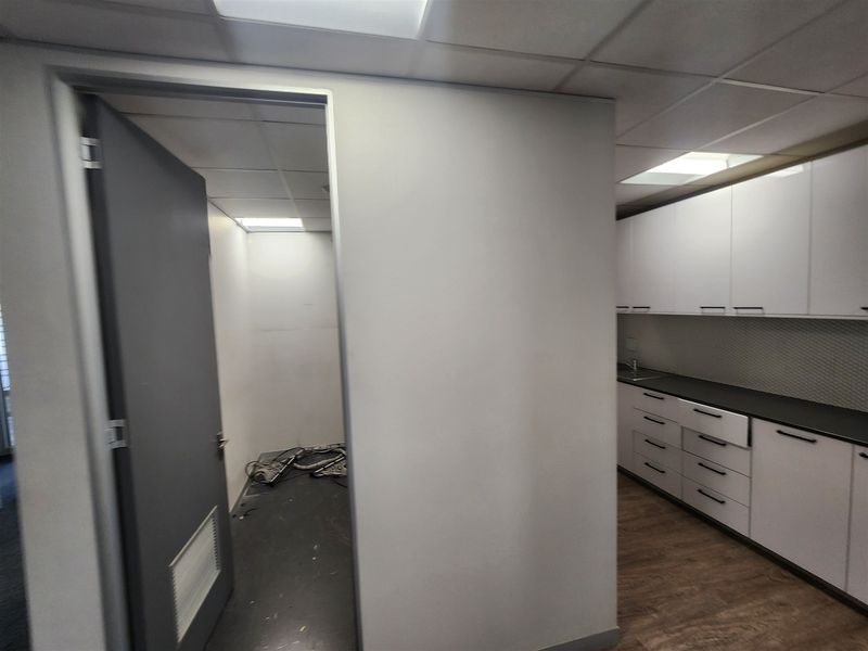 A functional office kitchenette featuring white cabinetry, dark countertops, and a drop ceiling, adjacent to an empty utility space with exposed wiring.
