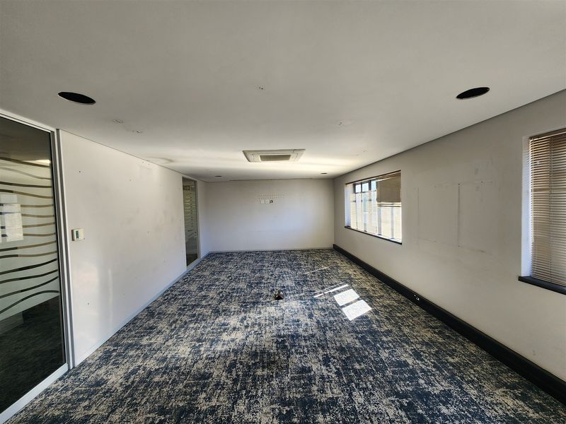 An empty office space featuring patterned carpeting, recessed ceiling lighting, a ducted air conditioning unit, and large windows with venetian blinds.
