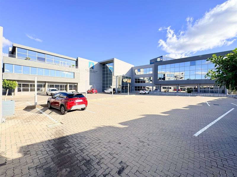 Modern multi-story office building featuring a glass facade and extensive paved parking area at the front entrance.