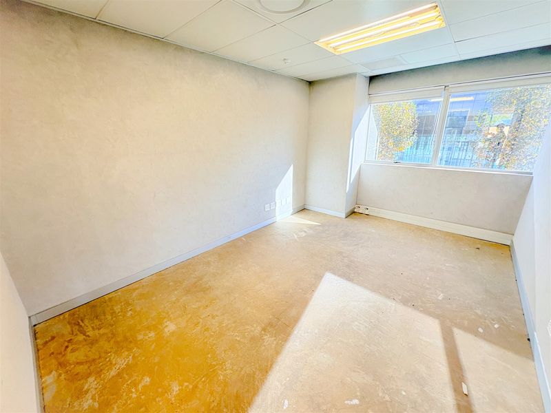 An empty office space featuring a suspended ceiling, fluorescent lighting, and a large window, currently requiring new flooring and wall finishes.