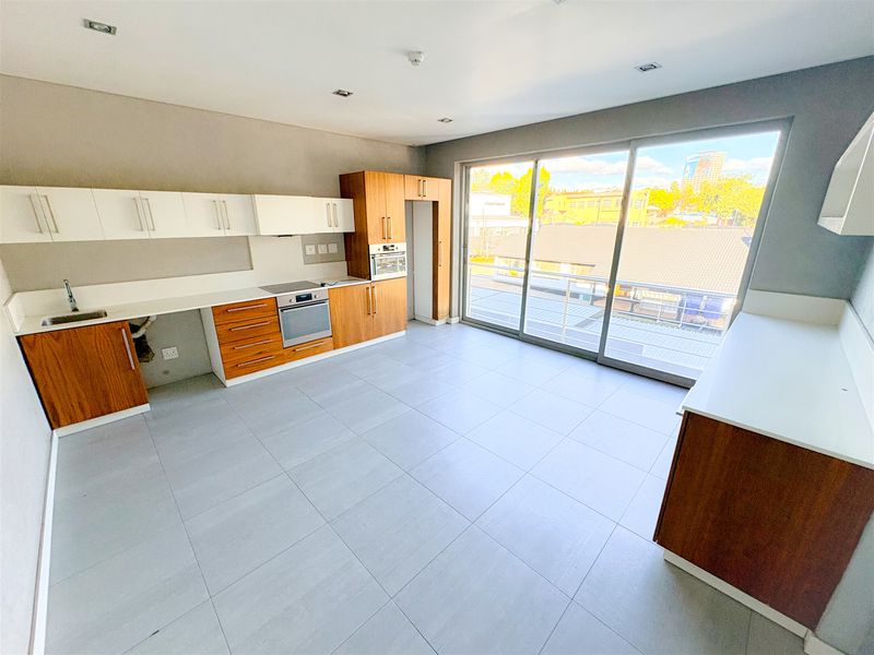Modern open-plan office kitchen area featuring wood-finish cabinetry, integrated appliances, tiled flooring, and large sliding doors leading to an outdoor balcony.