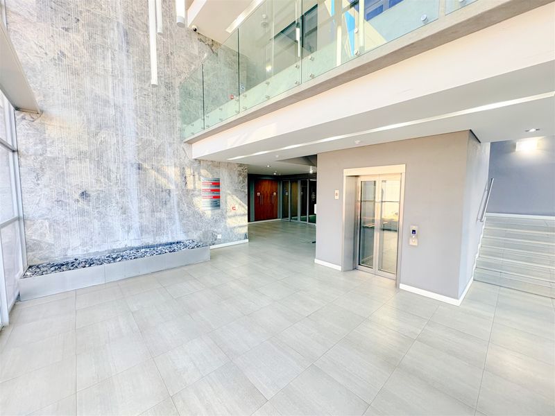 Modern office building entrance lobby featuring a striking stone water feature, glass balustrade, elevator access, and clean tiled flooring.