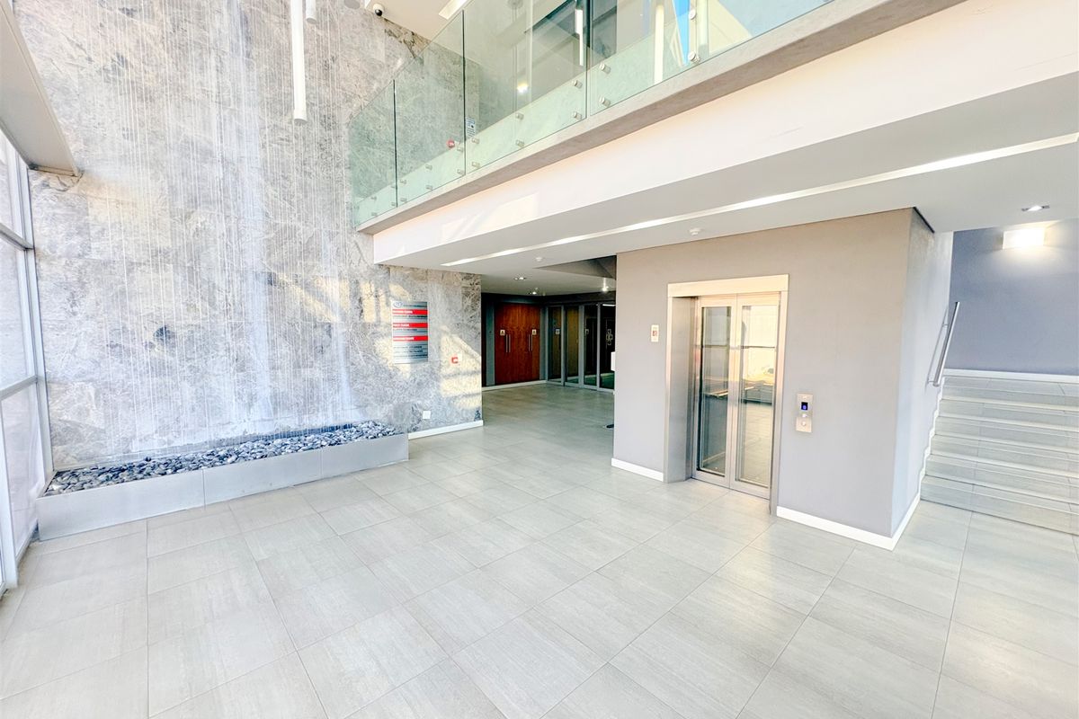 Modern office building entrance lobby featuring a striking stone water feature, glass balustrade, elevator access, and clean tiled flooring.