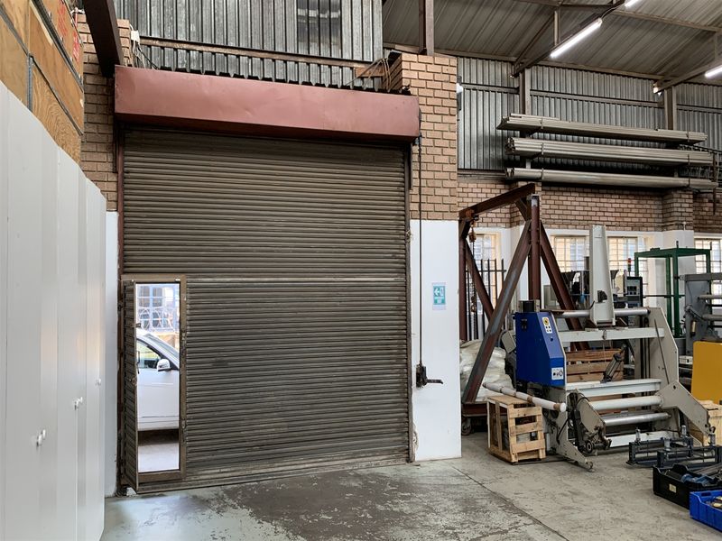 Industrial warehouse space featuring a large roller shutter door, exposed brick walls, high metal roof structure, and durable concrete flooring.