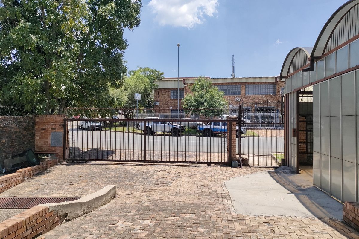 Front exterior view of the industrial property featuring a secure metal gate, brick paved driveway, and perimeter security fencing.