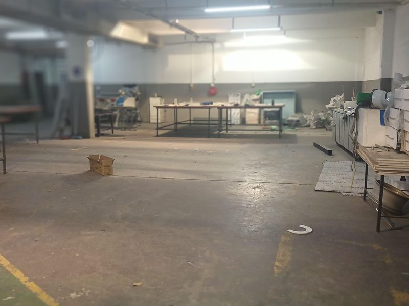 Spacious industrial workshop area featuring high ceilings, fluorescent lighting, and durable concrete flooring, suitable for manufacturing or storage operations.