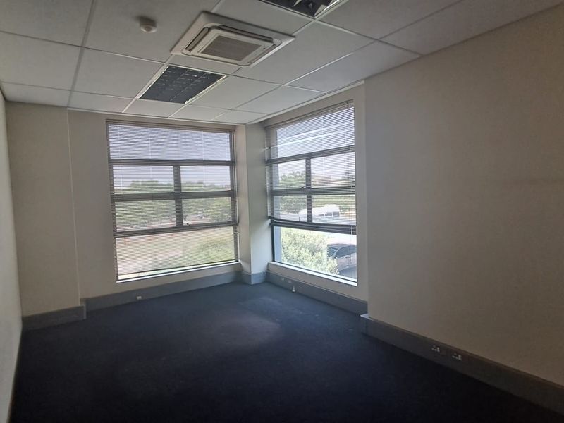 A vacant office space featuring large windows with integrated blinds, recessed ceiling lighting, and dark carpeting, suitable for a professional workspace.