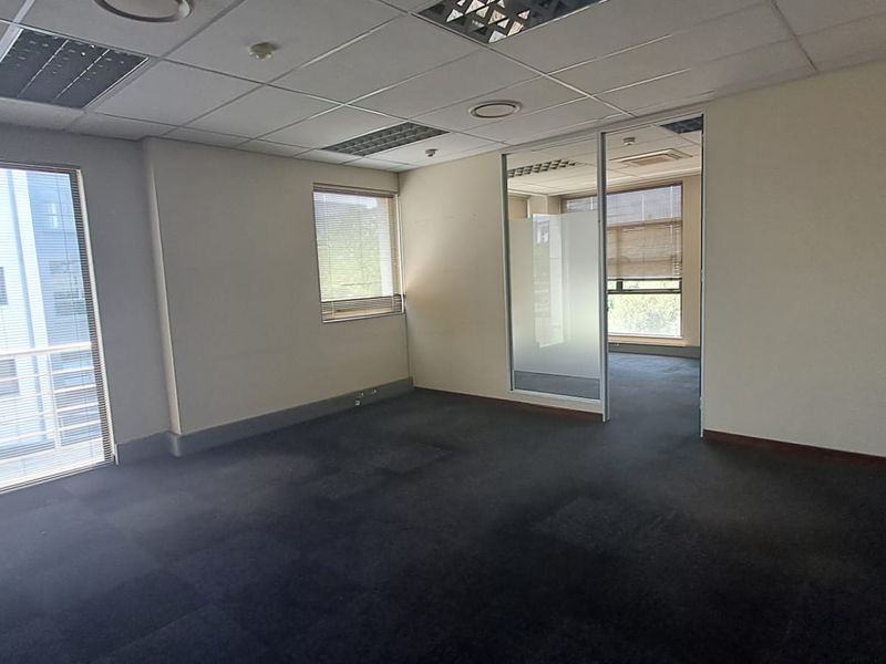 An empty office space featuring a suspended ceiling, carpeted flooring, and a glass partition wall leading to an adjacent office area.