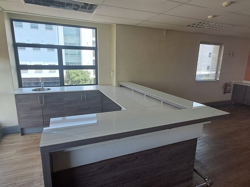 A modern office kitchenette featuring wood-grain cabinetry, a white countertop, and a built-in sink, set within a bright, open-plan workspace.