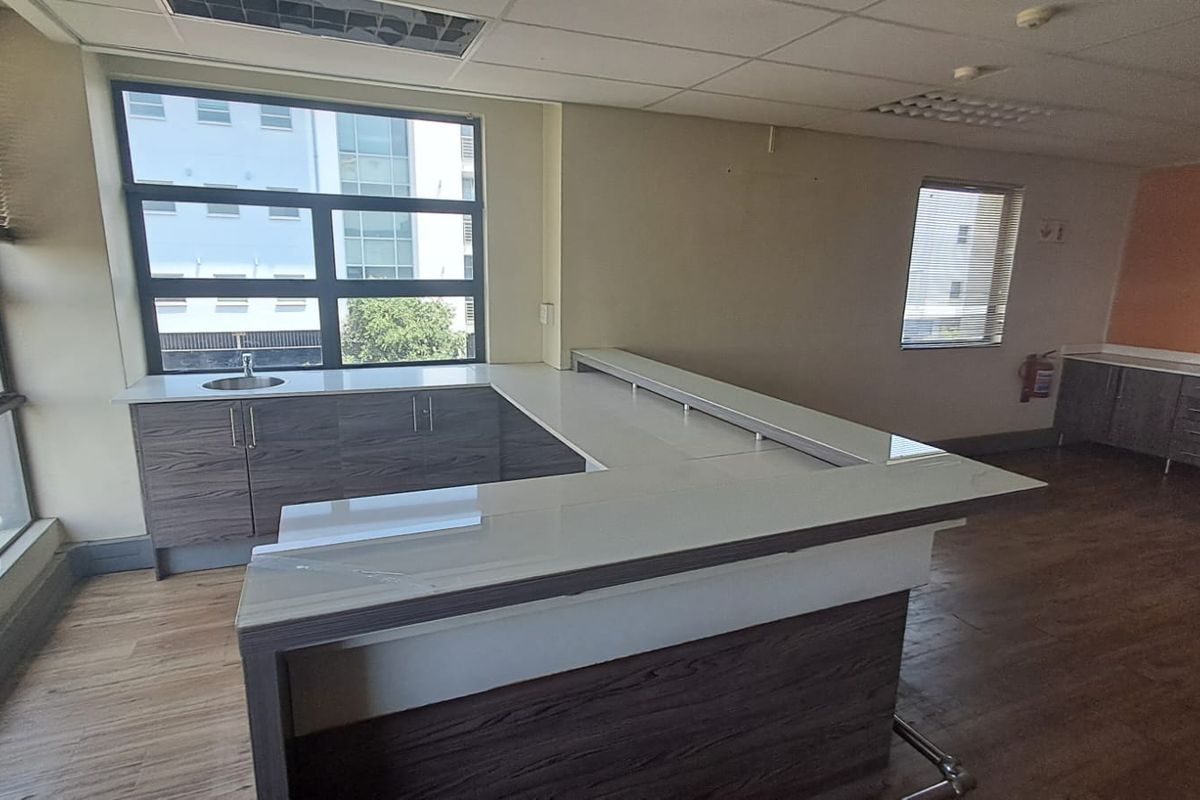 A modern office kitchenette featuring wood-grain cabinetry, a white countertop, and a built-in sink, set within a bright, open-plan workspace.