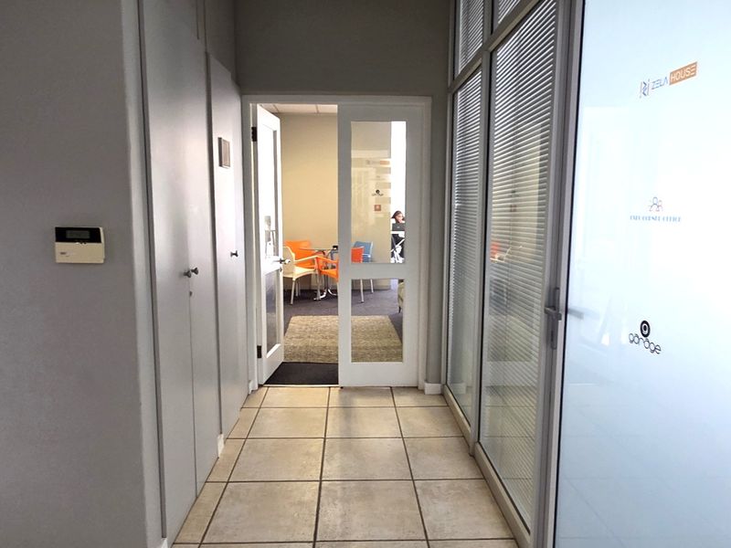 A clean office hallway featuring tiled flooring, built-in storage cupboards, and a glass partition wall leading to an adjacent office space.