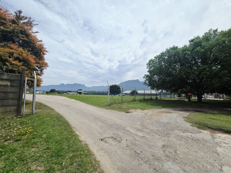 A view of the vacant commercial land featuring a gravel driveway, perimeter chain-link fencing, and mature trees with mountain views in the background.
