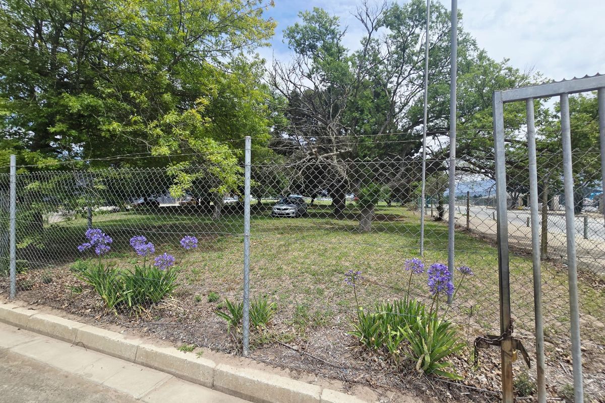 A view of a vacant commercial plot enclosed by chain-link fencing and a metal gate, featuring mature trees and established landscaping.
