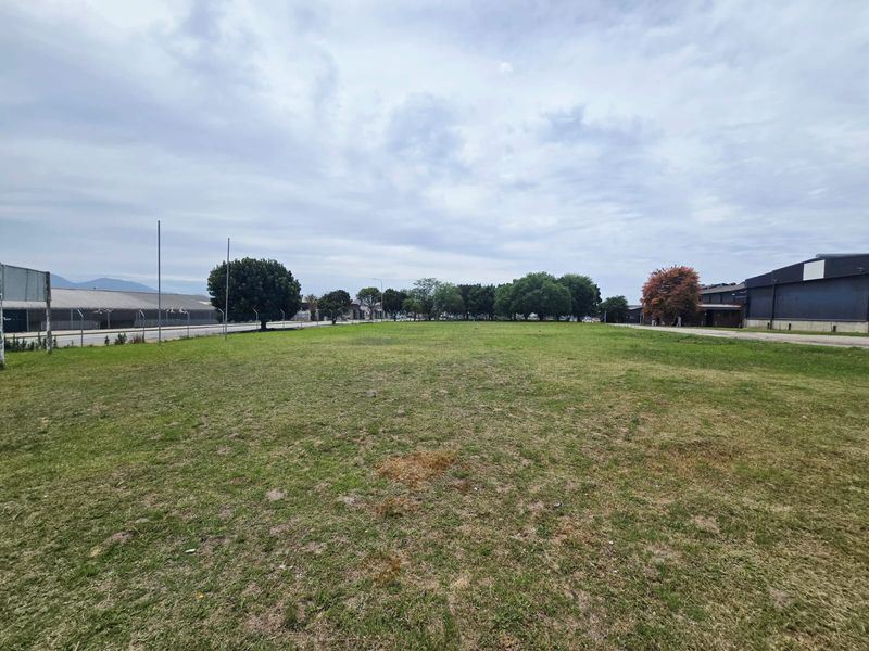 A wide, open vacant commercial lot featuring a grass lawn, perimeter chain-link fencing, and surrounding mature trees in a light industrial area.