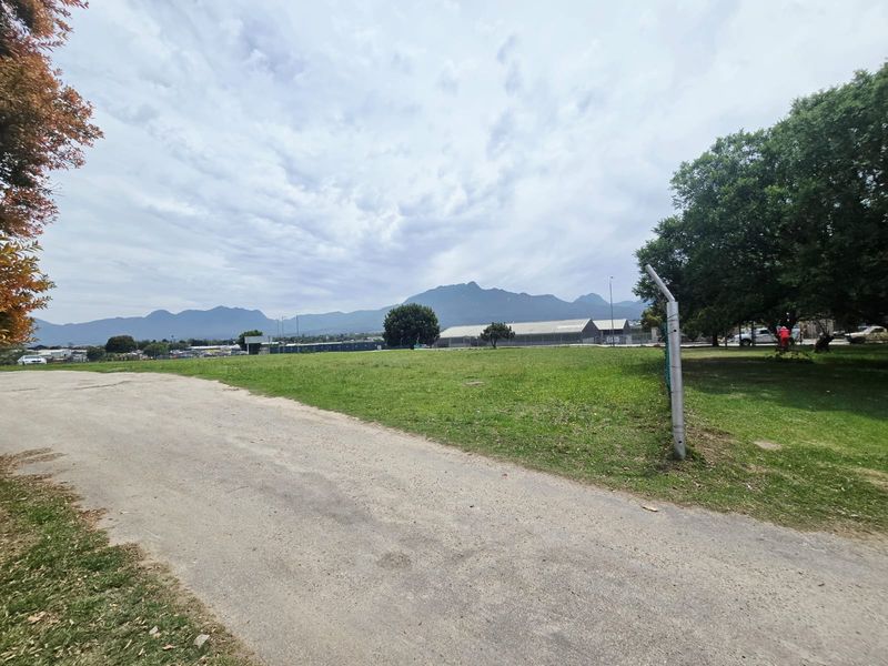 A wide-angle view of a vacant commercial land plot featuring a grass lawn, unpaved access path, and perimeter fencing with mountain views in the background.