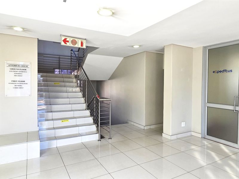 A clean and professional office building entrance lobby featuring tiled flooring, a central staircase, and glass-paneled office access doors.