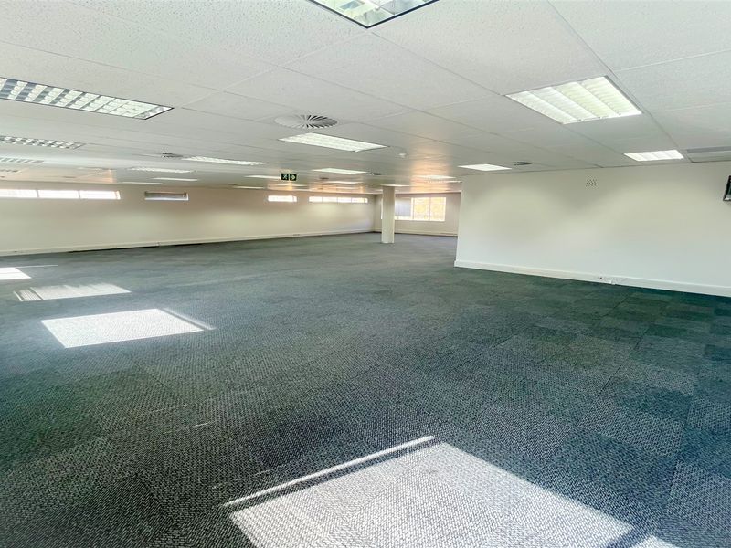 A spacious, open-plan office area featuring carpet tiles, a suspended ceiling with recessed lighting, and large windows providing natural light.