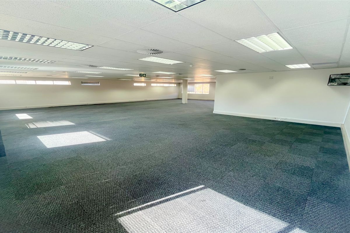 A spacious, open-plan office area featuring carpet tiles, a suspended ceiling with recessed lighting, and large windows providing natural light.