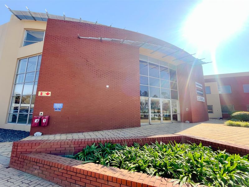 Front exterior view of a modern office building featuring a prominent red brick facade, large glass windows, and a paved entrance area.