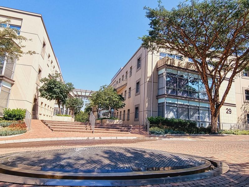A professional office park exterior featuring modern multi-story buildings with glass facades, a central paved courtyard, and a decorative water feature.