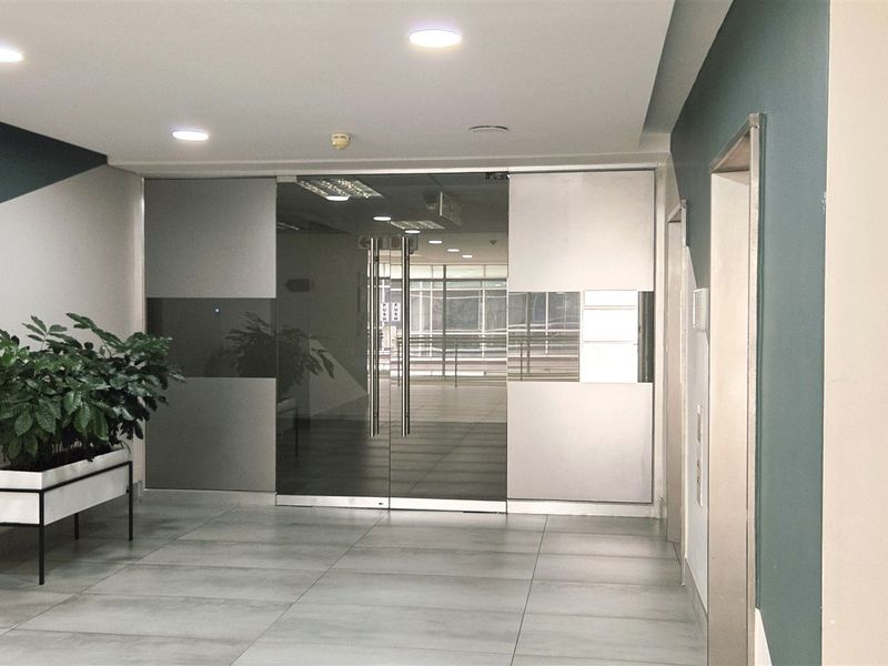Modern office building entrance lobby featuring sleek glass double doors, neutral tiled flooring, and contemporary recessed ceiling lighting.