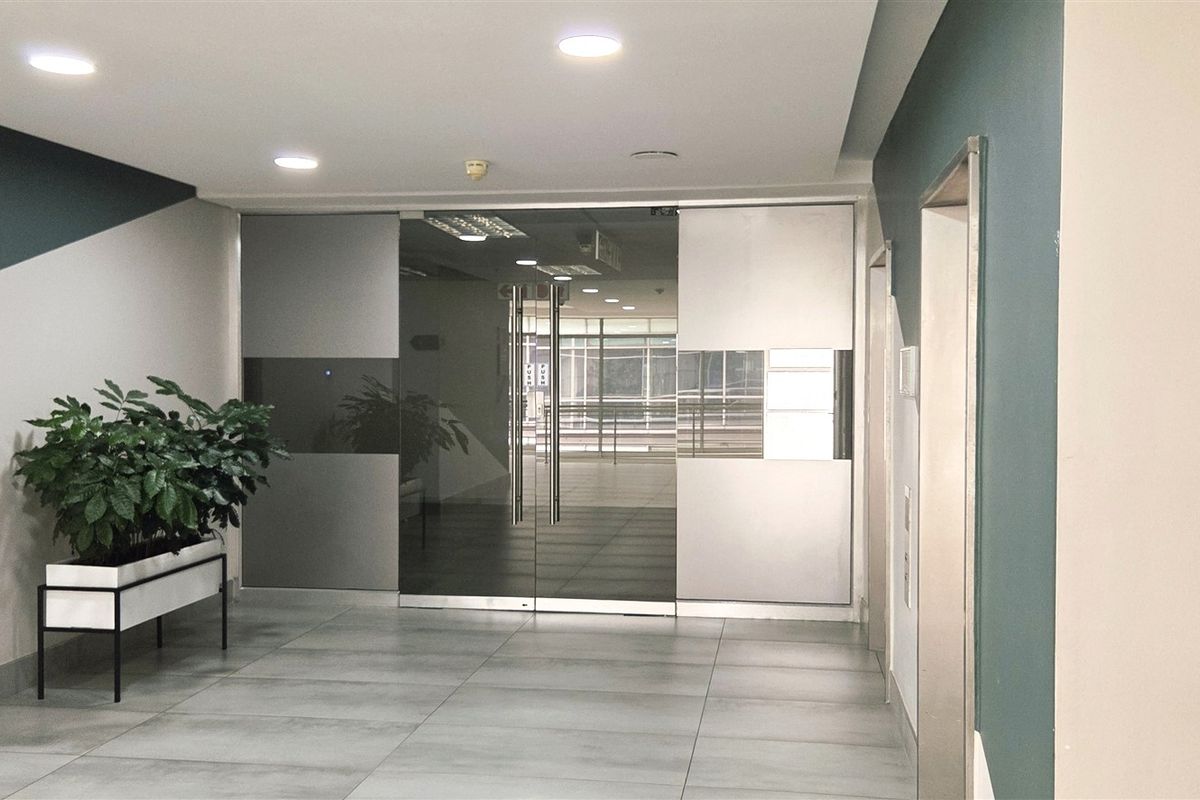 Modern office building entrance lobby featuring sleek glass double doors, neutral tiled flooring, and contemporary recessed ceiling lighting.