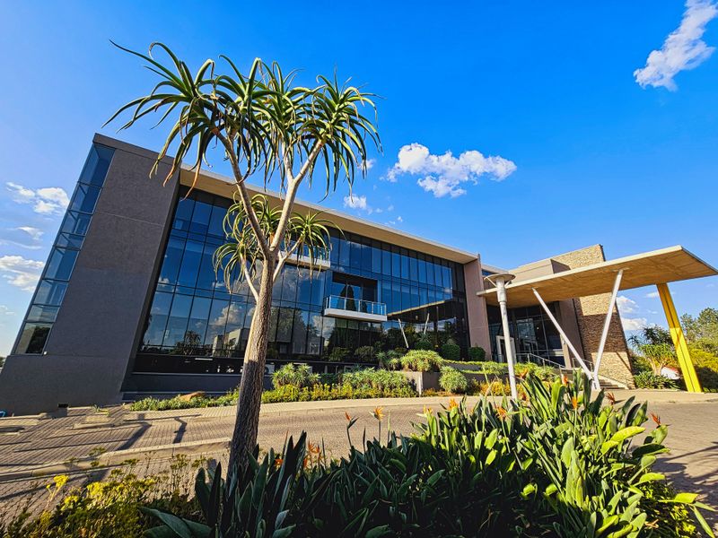 Modern office building exterior featuring a large glass facade, stone accents, and a prominent concrete entrance canopy with surrounding landscaped gardens.