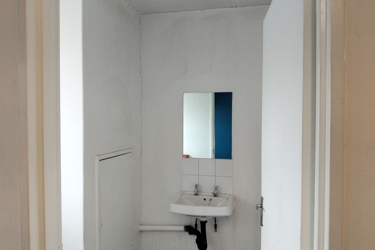 A small, functional bathroom space featuring a wall-mounted basin with tiled splashback and mirror, requiring cosmetic updates to improve the overall finish.